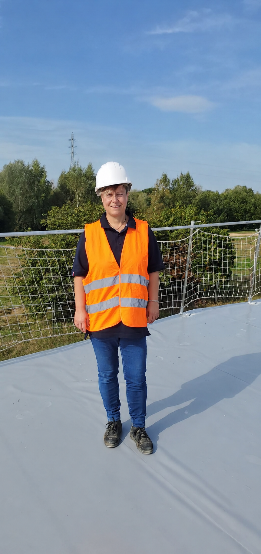 Virginie-Jacquemin-Experte-Bâtiment-PACA Portrait de Virginie Jacquemin, experte bâtiment indépendante en région PACA, portant un casque de chantier blanc et un gilet de sécurité orange sur un site en construction.